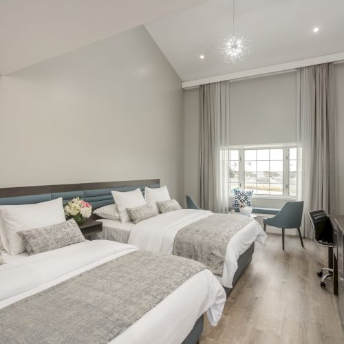 A modern hotel room with two large beds, a wall-mounted TV, a desk, a chair by the window, and minimalist decor with neutral tones.