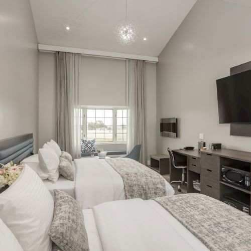 This image shows a modern hotel room with two beds, a flat-screen TV, a desk, a window, and contemporary decor.