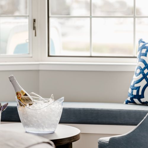 A cozy seating area with blue pillows, a large window, a chair, and an ice bucket with a bottle and flowers on a small table sits by the window.