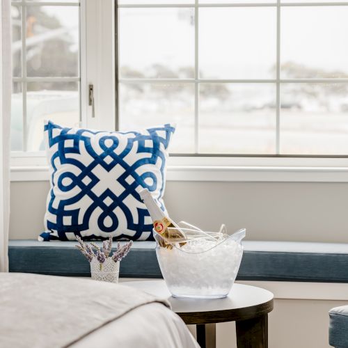 The image shows a cozy room with a decorative blue and white pillow, a window seat, a bucket of ice with champagne, and a side table with glasses.