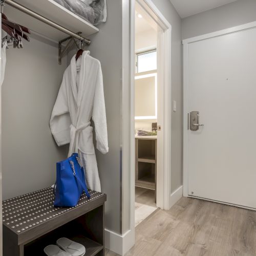 A hallway with a white door to the right, a closet with a bathrobe, hangers, and a blue bag on a shelf. Wood flooring throughout the space.