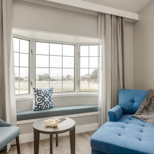 The image shows a cozy seating area with a blue chaise lounge, a matching chair, and a round table in front of a window with white curtains.