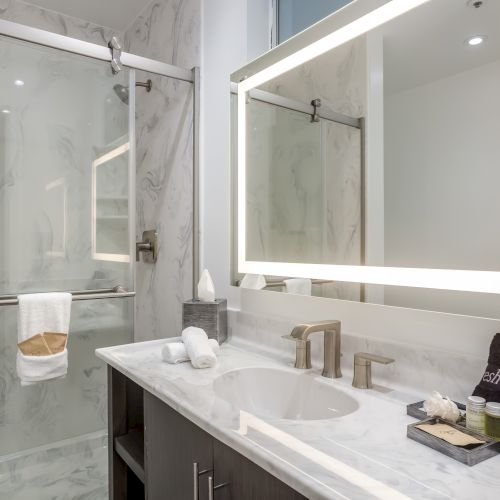 The image shows a modern bathroom with a marble countertop, illuminated mirror, glass shower door, and a neatly arranged set of amenities on the counter.