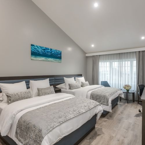 A modern hotel room with two large beds, a desk, a TV, and a window with curtains. Art on the wall depicts an ocean scene, and the room has wooden floors.