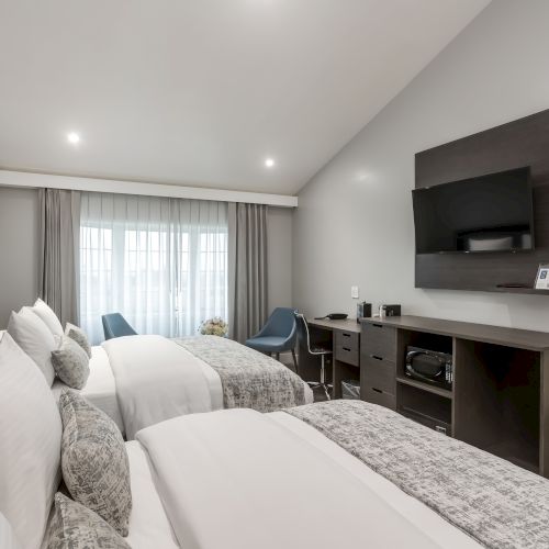 A hotel room with two neatly made beds, a flat-screen TV, a desk, chairs, and large window with curtains.