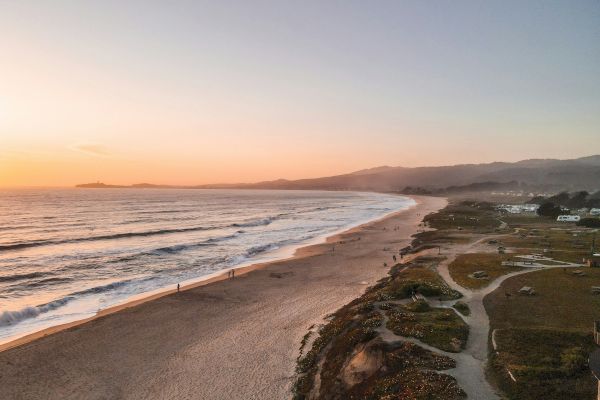 A serene beach scene at sunset with gentle waves, a sandy shore, and distant mountains under a colorful sky in the background.