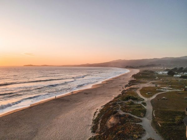 A serene beach scene at sunset with gentle waves, a sandy shore, and distant mountains under a colorful sky in the background.