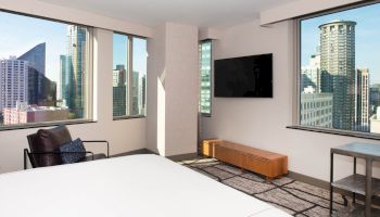 The image shows a modern hotel room with a bed, chair, TV, and large windows displaying a city view with tall buildings.