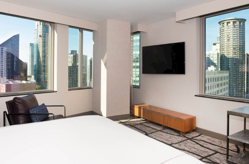 The image shows a modern hotel room with a bed, chair, TV, and large windows displaying a city view with tall buildings.