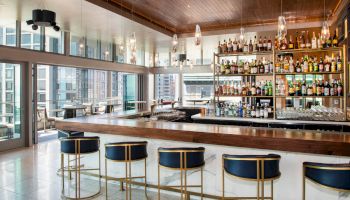 A stylish bar with a fully stocked shelf, modern decor, blue and gold bar stools, large windows with a city view, and an inviting atmosphere.