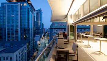 A modern city view at dusk from an open, stylish balcony featuring outdoor seating and tables, overlooking high-rise buildings and a street below.