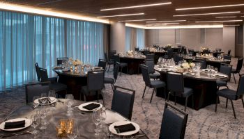 The image shows an elegantly set banquet hall with round tables, black chairs, and white floral centerpieces. The room has large windows with blue curtains.
