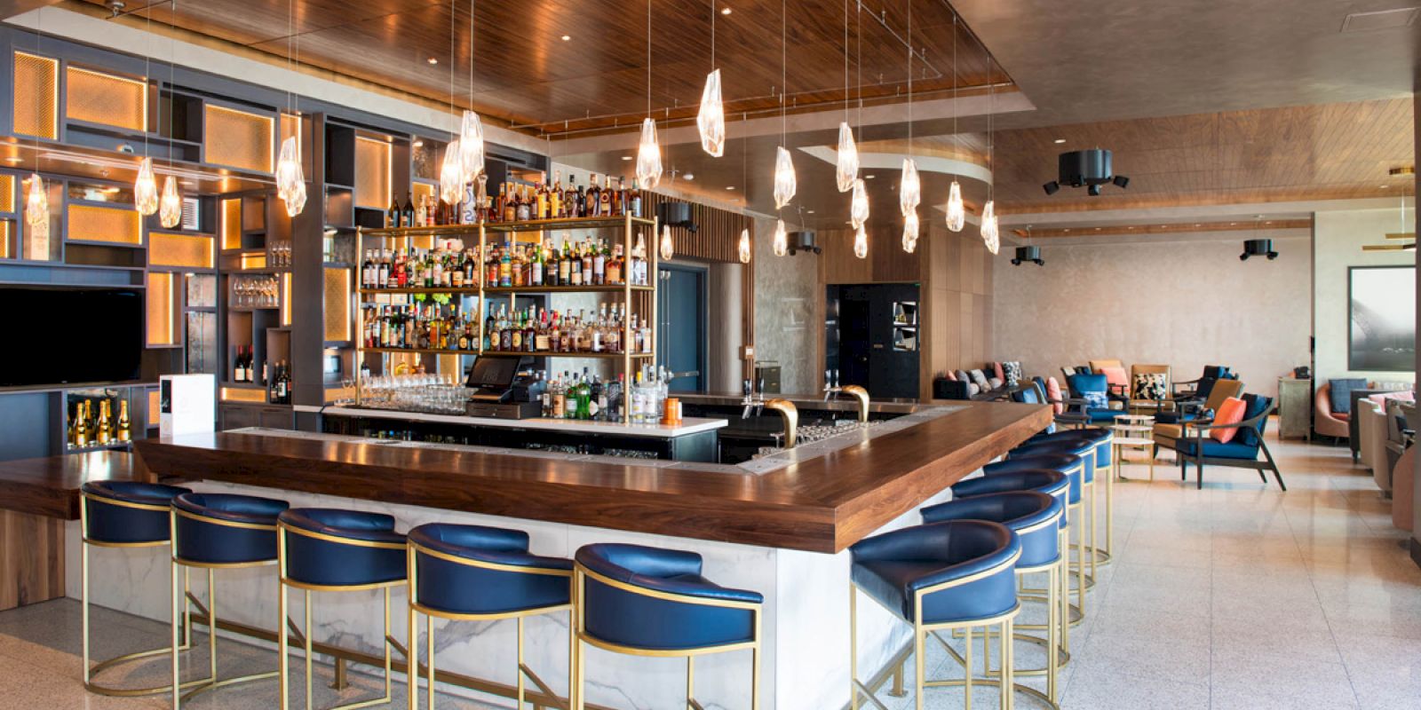A modern bar with blue cushioned stools, a stocked shelf of bottles, pendant lights, and contemporary furniture in a spacious, well-lit setting.