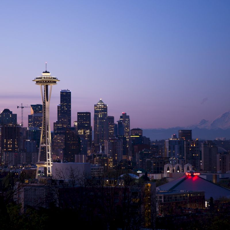 The image shows a dusk view of a city skyline featuring the Space Needle with Mount Rainier in the background, under a clear sky.