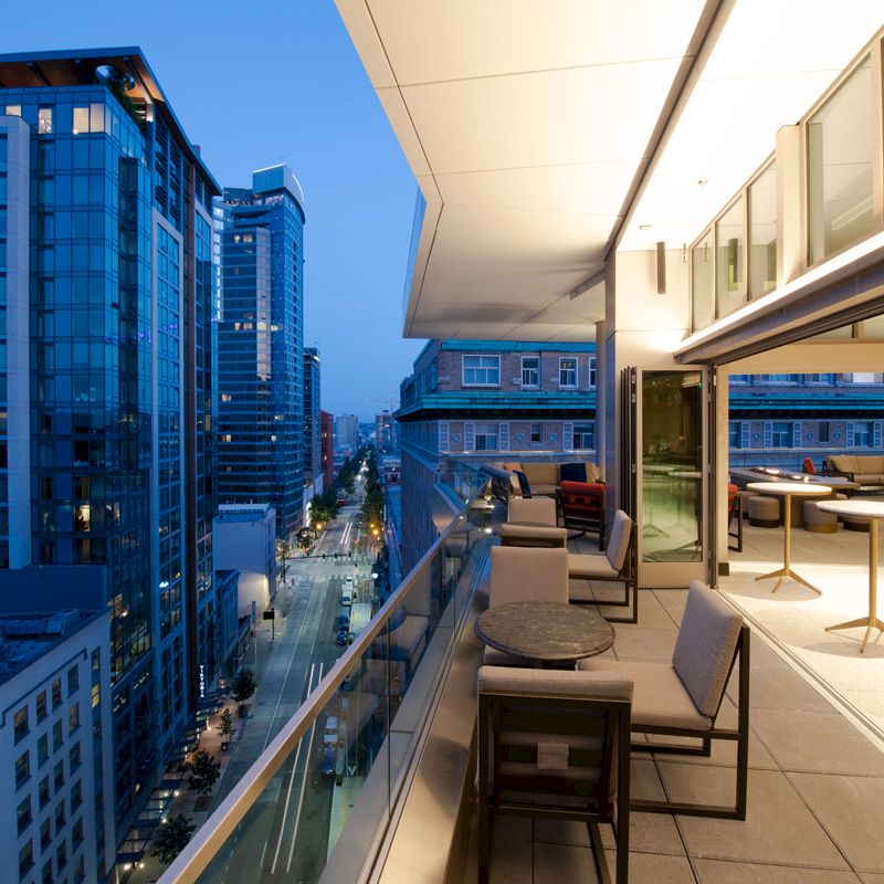 The image shows a modern, well-lit outdoor terrace with seating, overlooking a cityscape with tall buildings during dusk or evening.