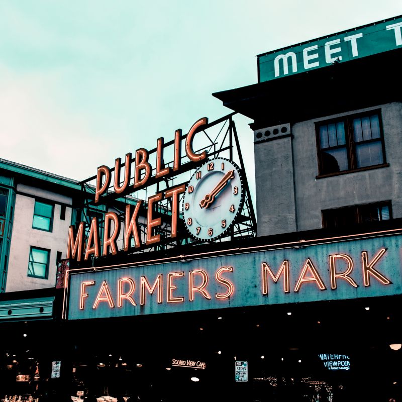 The image depicts an iconic public market with a prominent clock sign, and a neon sign reading "FARMERS MARKET" in a bustling urban setting.