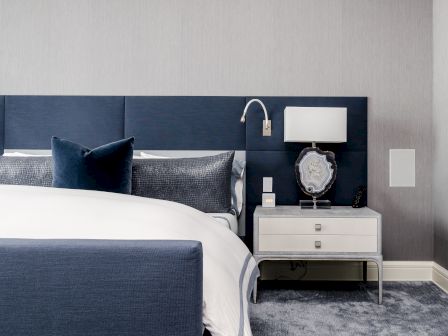 A modern bedroom with a blue and white color scheme, including a blue upholstered headboard, a nightstand with a lamp, and decorative pillows.