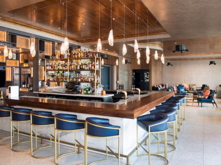 The image shows a modern bar with blue barstools, a well-stocked bar, hanging pendant lights, and a seating area in a stylish, contemporary design.