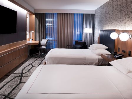 The image shows a modern hotel room with two beds, a wall-mounted TV, a workspace, and stylish decor with wallpaper and curtains.