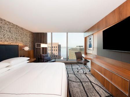 The image shows a modern hotel room with a bed, large TV, desk, chair, wall art, and a window view of buildings and water in the background.