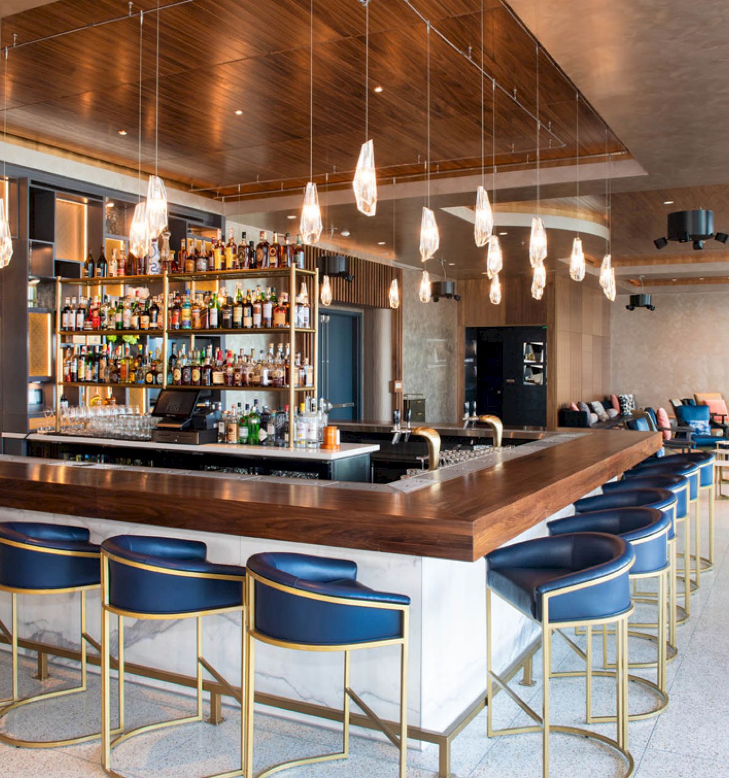 This image shows a modern bar with blue chairs, hanging lights, and a well-stocked liquor shelf. There's a seating area in the background.