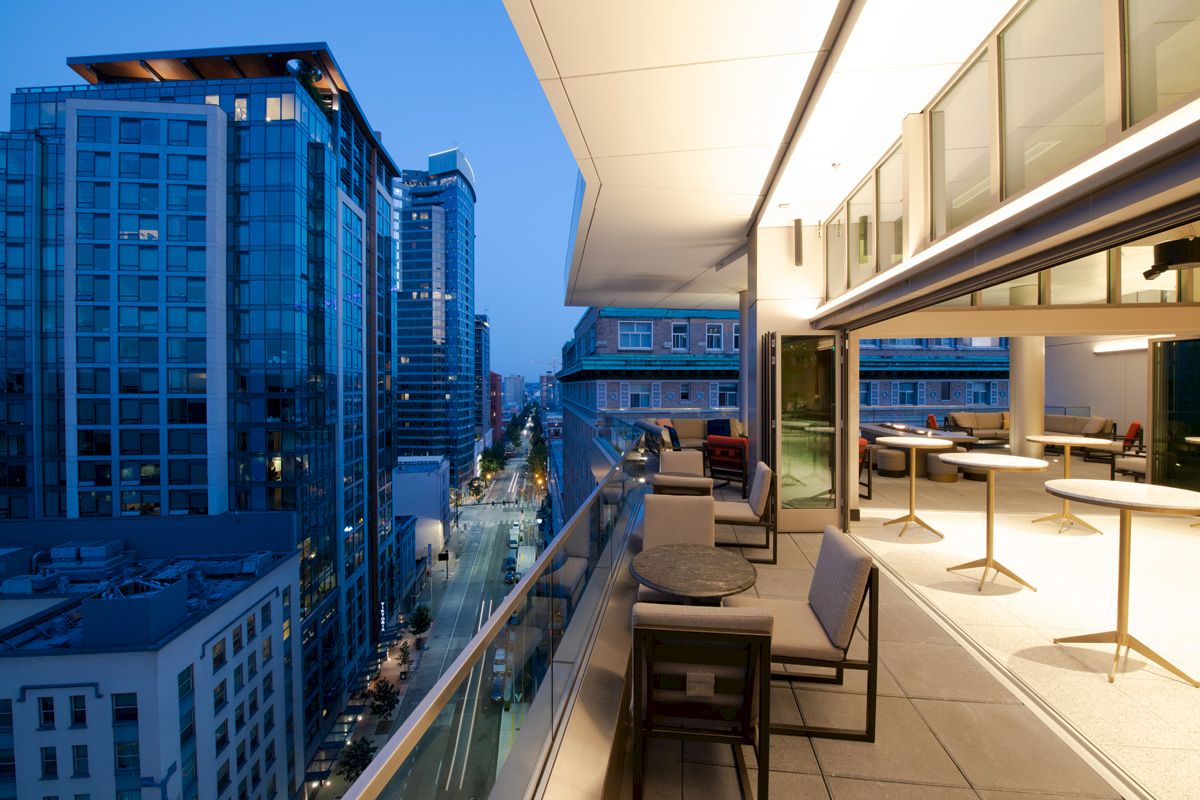 The image shows a modern outdoor balcony at dusk with a view of tall city buildings, furnished with tables and chairs, and well-lit interior spaces.