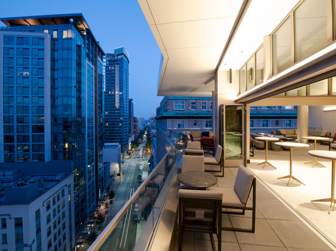 The image shows a modern outdoor balcony at dusk with a view of tall city buildings, furnished with tables and chairs, and well-lit interior spaces.