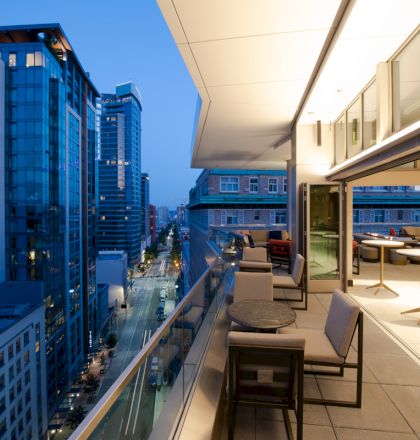 The image shows a modern urban balcony with sleek furniture, overlooking a street between tall buildings during twilight.