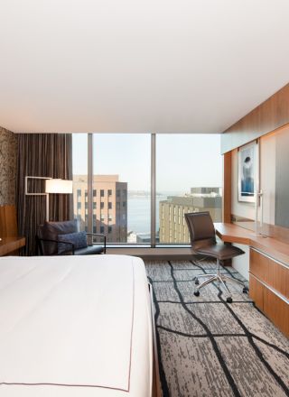 A modern hotel room with a king-sized bed, flat-screen TV, desk, chair, and large window offering a city view.