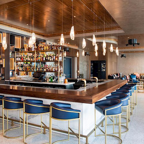 A modern bar with high stools, a well-stocked liquor shelf, pendant lights, and a seating area with sofas in the background.