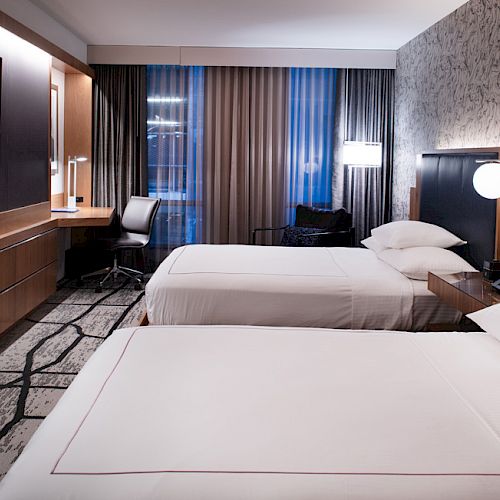 The image shows a modern hotel room with two neatly made beds, a wall-mounted TV, a study desk with a chair, and stylish lighting and decor.