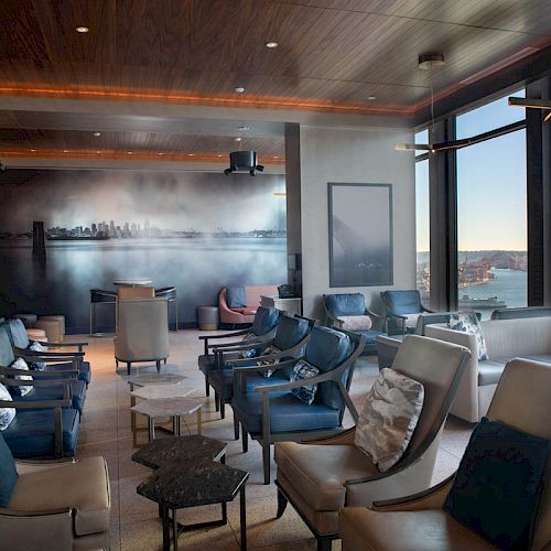 A modern lounge with comfortable seating, a cityscape mural, large windows offering a scenic view of a bay, and a Ferris wheel in the distance.