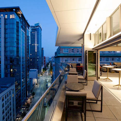 A modern balcony overlooking a cityscape at dusk, with outdoor seating and tables. The interior is visible through large sliding glass doors.