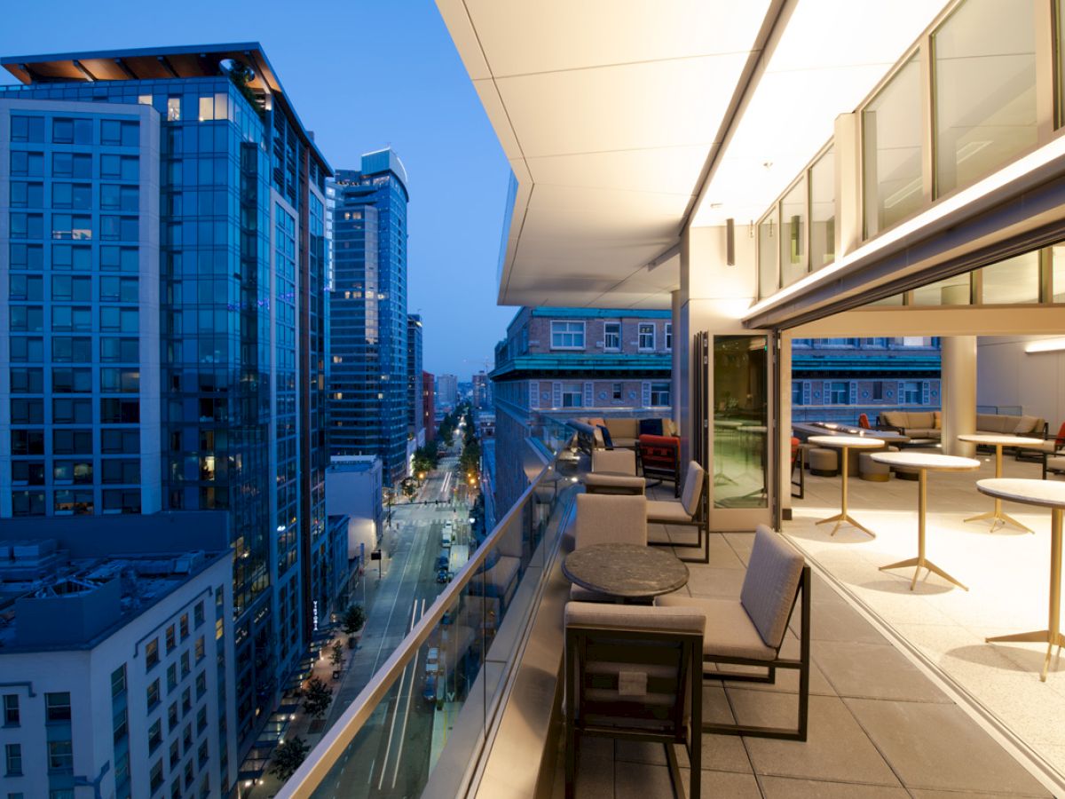 This image shows a modern balcony with seating and tables overlooking an urban street with tall buildings at dusk.