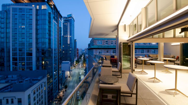 This image shows a modern balcony with seating and tables overlooking an urban street with tall buildings at dusk.