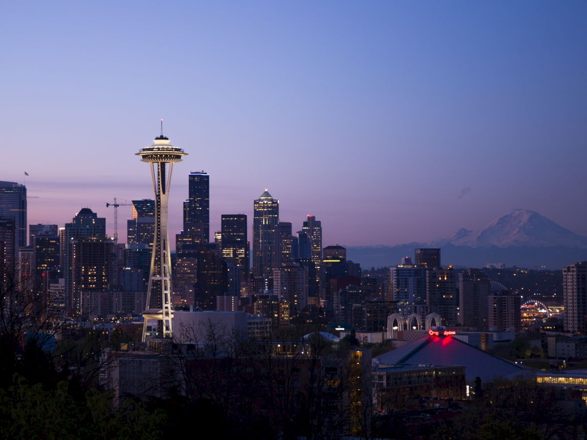 A cityscape at dusk featuring a skyline with a prominent tower and a distant mountain under a colorful sky.