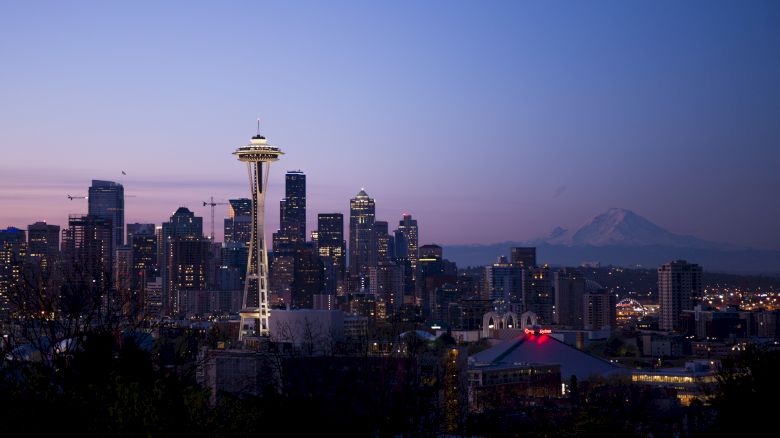 A cityscape at dusk featuring a skyline with a prominent tower and a distant mountain under a colorful sky.