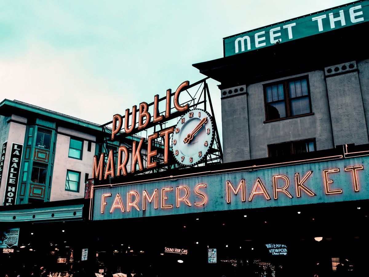 The image shows an outdoor public market with a neon sign reading "Public Market" and "Farmers Market," featuring a clock and surrounding buildings.