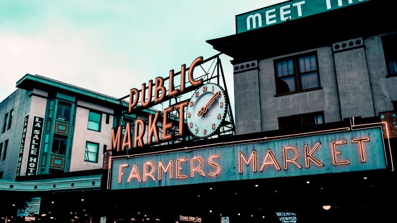 The image shows an outdoor public market with a neon sign reading "Public Market" and "Farmers Market," featuring a clock and surrounding buildings.