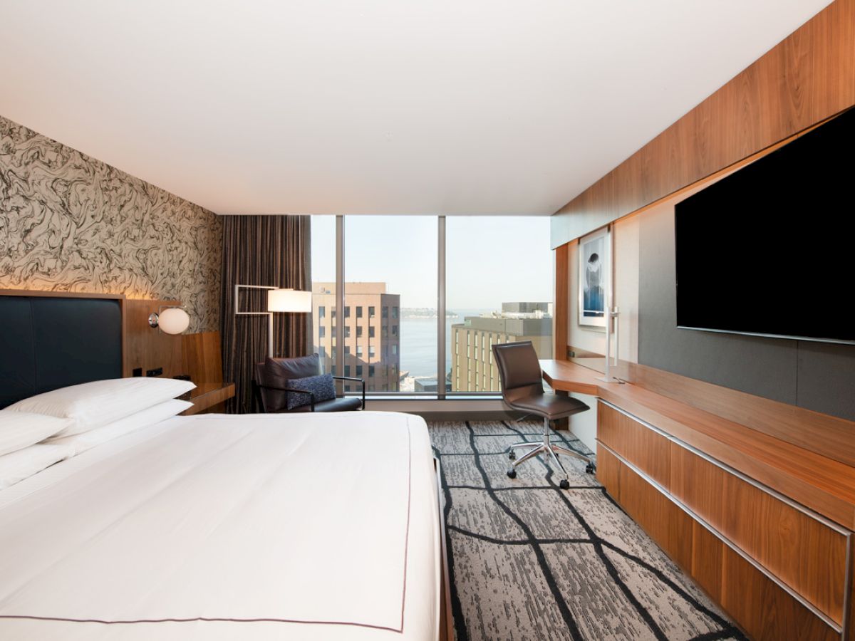 A modern hotel room with a large bed, a wall-mounted TV, a desk and chair, and a window offering a cityscape view.
