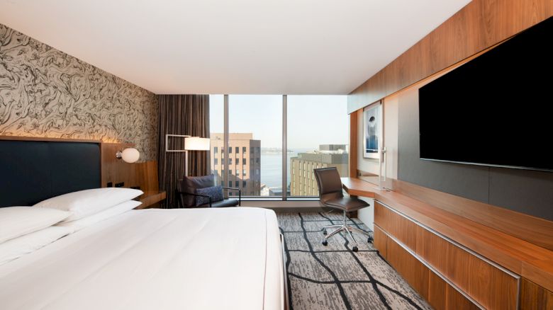 A modern hotel room with a large bed, a wall-mounted TV, a desk and chair, and a window offering a cityscape view.