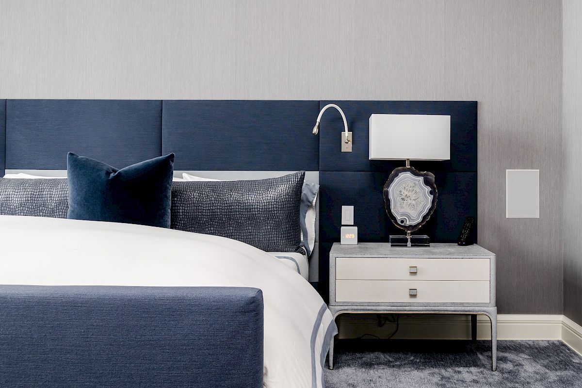 A modern bedroom with a blue and white color scheme; features a bed with blue headboard, pillows, and a nightstand with a lamp and decor.