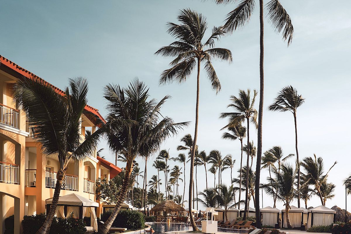 An outdoor pool area with lounge chairs, palm trees, and a building with balconies against a clear sky is shown in the image.