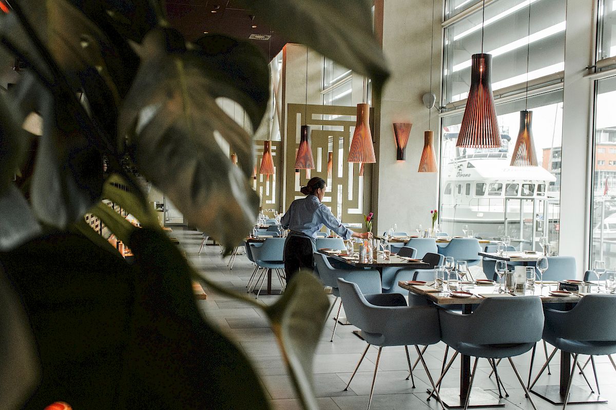 The image shows a restaurant interior with a staff member setting tables, modern decor, large windows, and distinctive light fixtures.
