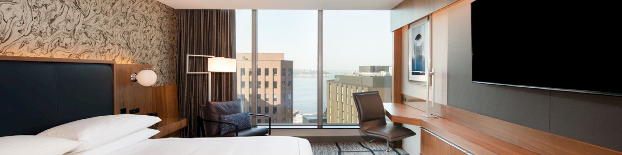 A modern hotel room with a large bed, wall-mounted TV, desk, ergonomic chair, and a window overlooking city buildings and water view.